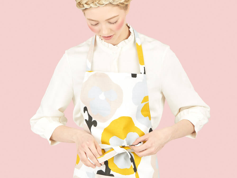 Woman wearing Kauniste´s apron with Orvokki pattern design, created by Hannele Äijälä